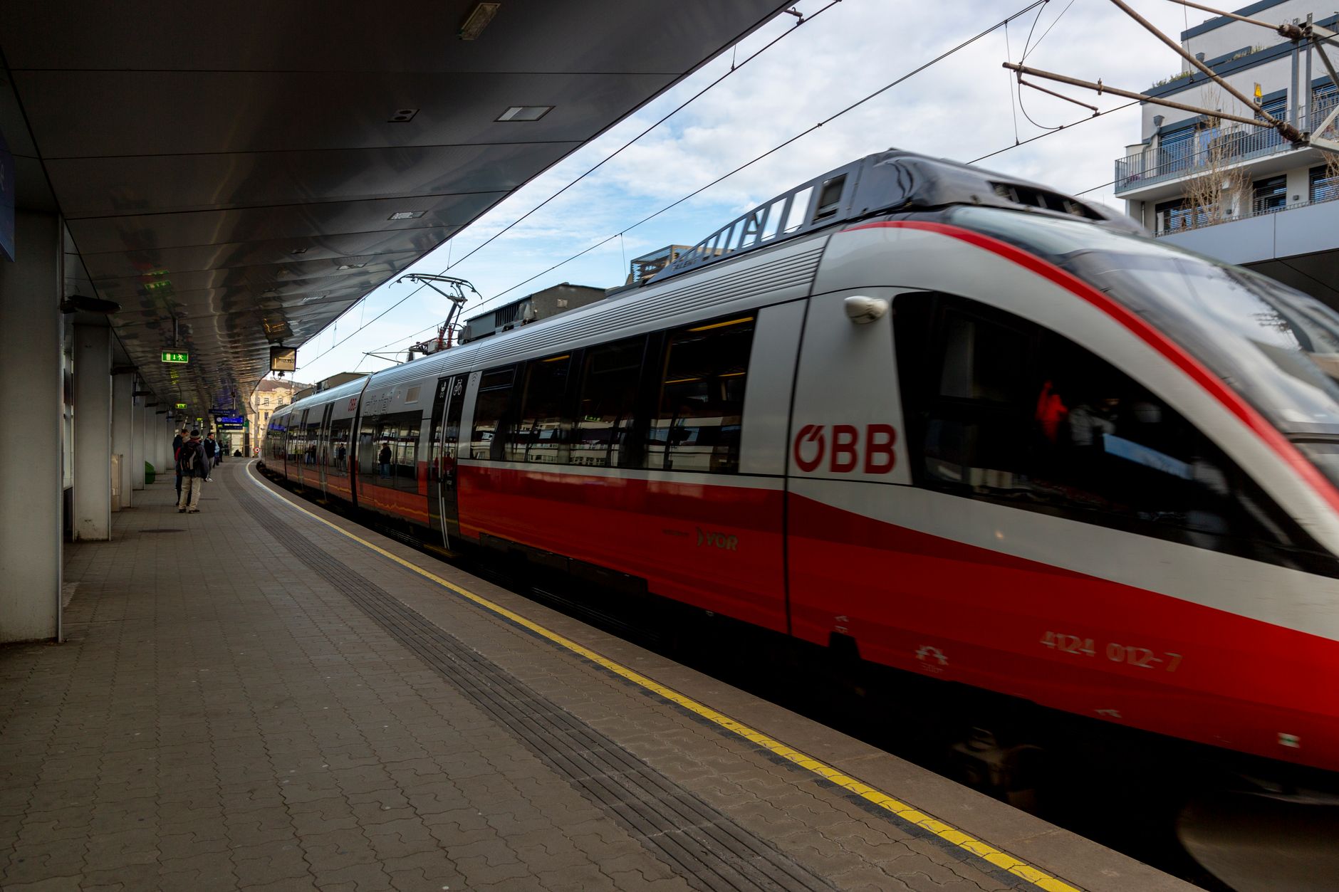 A sleek red and white ÖBB train on the platform at a modern station, with passengers waiting.