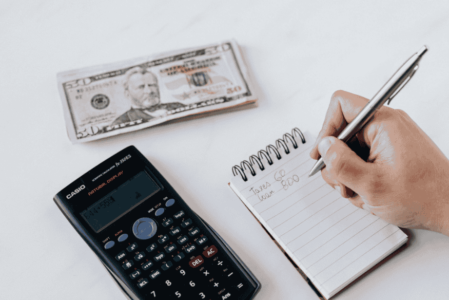 Person writing expenses in a notebook with calculator and cash to assess personal financial health