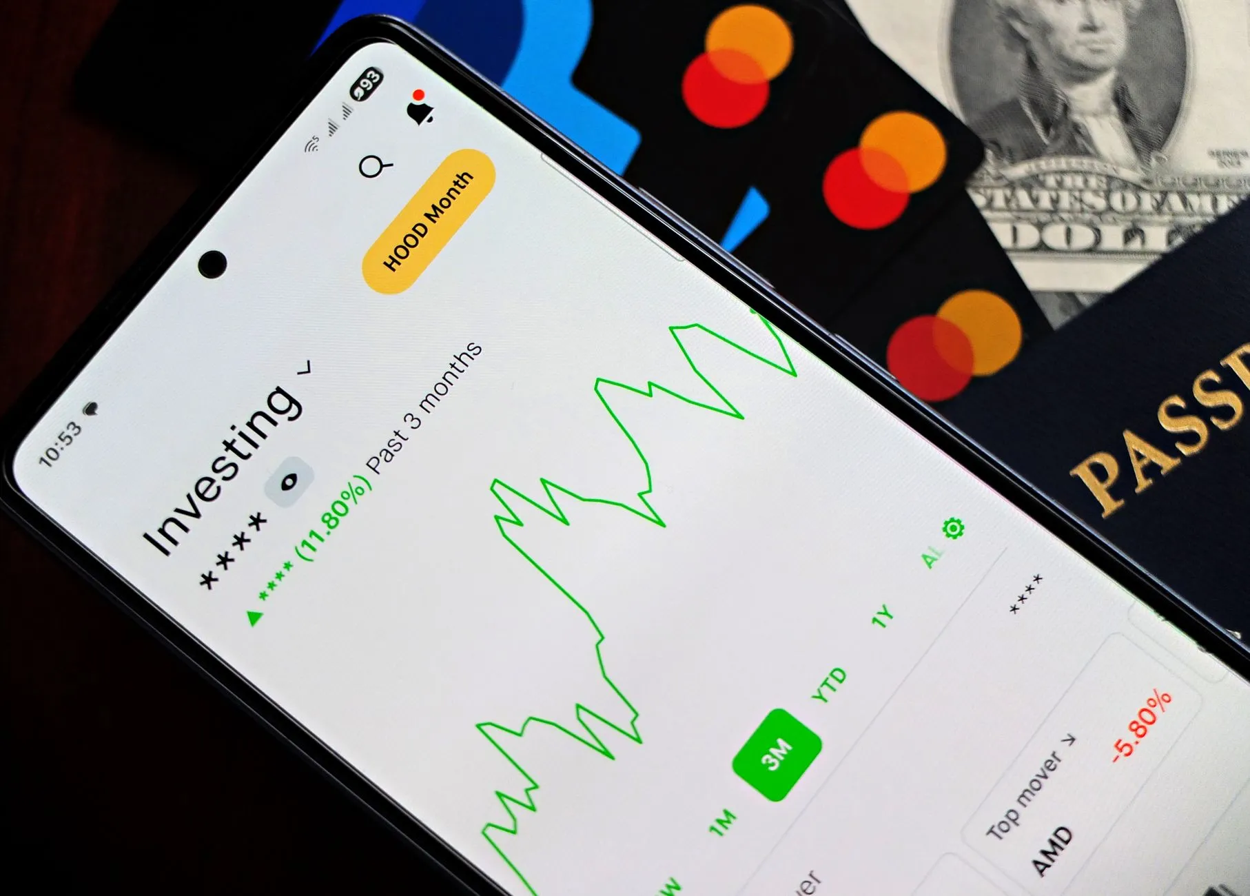 A smartphone showing an investment app with green growth indicators, surrounded by credit cards, US dollars, and a passport.