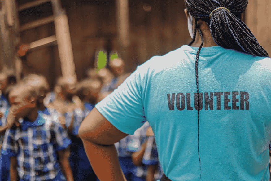 Mulher de costas com camisa de voluntária diante de grupo de crianças.