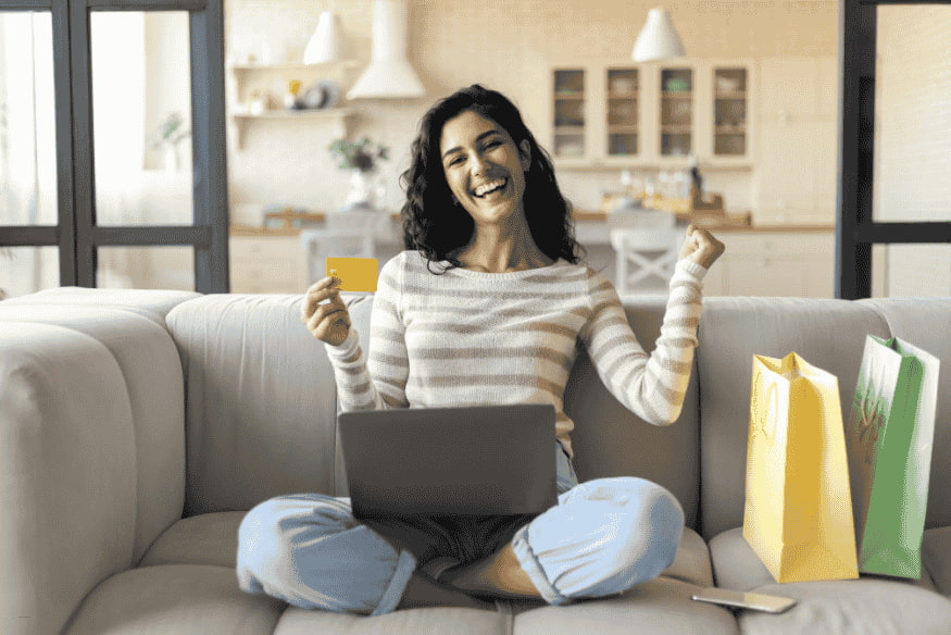Happy woman shopping online with a credit card and laptop, celebrating cashback rewards at home.