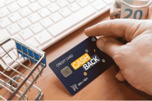 Hand holding a cashback credit card near a computer keyboard and shopping cart icon, symbolizing online rewards.