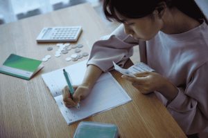 Young woman tracking her monthly expenses in a notebook with receipts, coins and a calculator on the table, illustrating simple expense tracking methods at home.