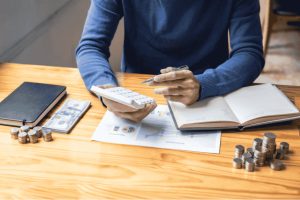 Person using a calculator with coins, cash and notebook on the table while creating a monthly budget to organize personal finances.