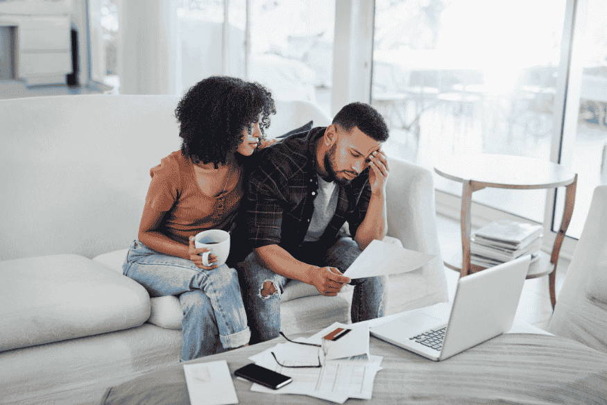 Worried couple on the couch reviewing bills and bank statements on a laptop, showing how overspending impacts household finances.